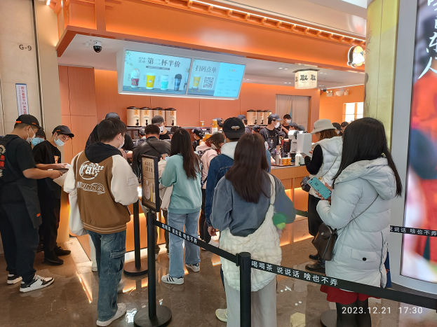 Eine Gruppe von Menschen steht in einer Schlange an einem Tresen in einem Restaurant. Einige tragen Mützen und tragen Taschen, während andere Gegenstände in den Händen halten. Es gibt Stangen mit Bändern und ein Display mit Text, Deckenleuchten und Tische mit verschiedenen Gegenständen im Hintergrund.