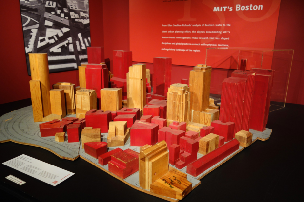 Ein hölzernes Blockmodell einer Stadt auf einem Tisch in einem Museum, begleitet von Papieren, einer Glasbox und einem Rahmenfoto an der Wand.