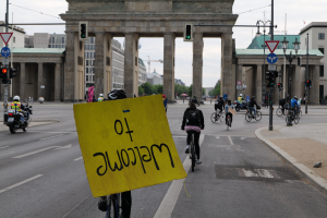 Eine Gruppe von Radfahrern mit Helmen fährt eine Straße entlang vor dem Brandenburger Tor in Berlin, Deutschland, mit einer Person, die ein gelbes Schild hält, Laternenpfählen, Verkehrszeichen, Gebäuden, Bäumen und einem klaren blauen Himmel im Hintergrund.