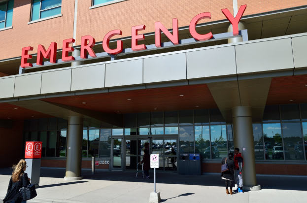 Eine Gruppe von Menschen vor einem Glaswand-Notfallgebäude mit Säulen, einem Namensschild, Hinweistafeln an Pfählen und einem Mülleimer.