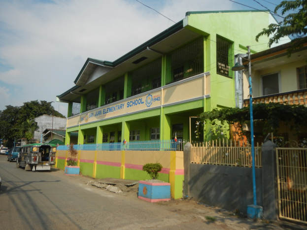 Schulgebäude mit grüner und gelber Fassade, umgeben von Fahrzeugen, Topfpflanzen, einem Zaun, einem Tor, einem Straßenschild, Bäumen, Drähten und einem bewölkten Himmel.