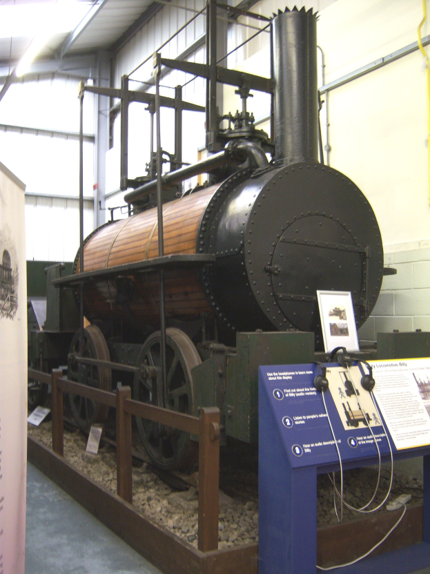 Alte Dampflokomotive in einem Museum mit Informationsschildern, einem Zaun, Steinen, einer Fahne, einer Wand mit Rohren und Deckenbeleuchtung.