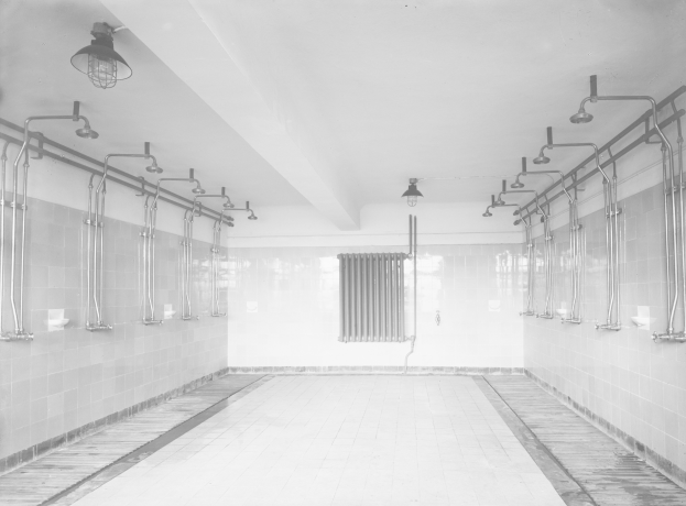 Schwarzes und weißes Foto einer öffentlichen Toilette mit Fliesenboden und Wänden, sichtbare Rohre, Lichter, Decke und verstreute Gegenstände.