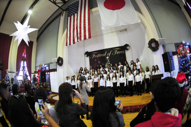 Gruppe von Menschen auf einer Bühne mit Weihnachtsbäumen, einem Rednerpult, Lautsprechern, einer Texttafel, Vorhängen, Fahnen und Deckenleuchten vor einem Publikum mit Kameras und Handys.