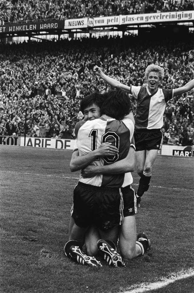 Drei Fussballspieler umarmen sich auf dem Boden beim Feiern eines Tors, mit jubelnder Menge und schwenkenden Fahnen im Hintergrund, dargestellt in Schwarz-Weiss.