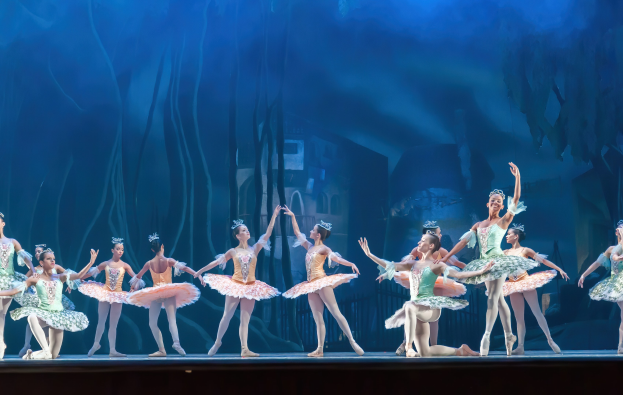 Eine Gruppe von Balletttänzern in farbenfrohen Kostümen, die *Der Nussknacker* auf der Bühne des Royal Opera House tanzen, synchronisiert gegen einen blauen Hintergrund.