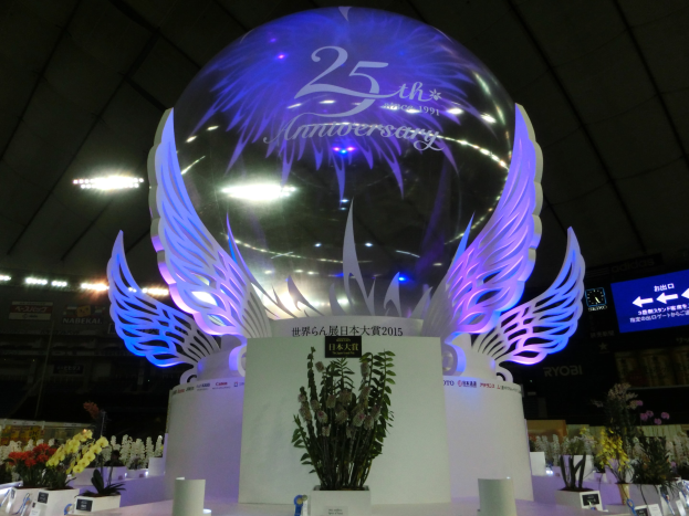 Ein großer Kristallball mit Flügeln oben drauf, umgeben von Blumenschmuck, Namensschildern und anderen Gegenständen auf dem Boden, vor einer Kulisse aus Brettern, Lichtern und einem Bildschirm während einer 25-jährigen Jubiläumsfeier.