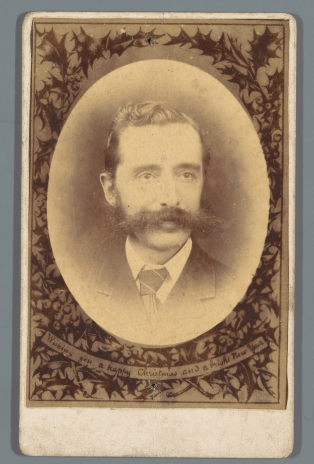 Ein altes Foto eines Mannes mit Schnurrbart und Bart, der einen Anzug und eine Krawatte trägt und direkt in die Kamera schaut.