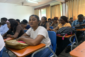Eine Gruppe von Menschen, die an Tischen in einem classroom der University of Ghana sitzt, mit Taschen und Gegenständen auf den Tischen, Fenstern mit Vorhängen, einer Klimaanlage an der Wand und Lampen und Ventilatoren an der Decke im Hintergrund.