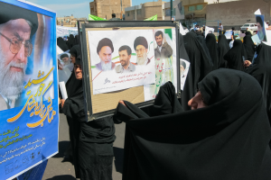 Eine Gruppe von Frauen in schwarzen Abayas hält Plakate mit Fotos von Ajatollah Ali Khamenei hoch, im Hintergrund sind Gebäude, Pfosten, Fahrzeuge und ein klarer blauer Himmel zu sehen.