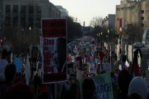 Menschen mit Plakaten auf einer Straße protestierend, mit Gebäuden, Bäumen, Laternen und Himmel im Hintergrund.