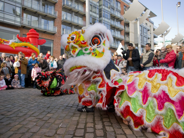 Ein farbenfrohes chinesisches Neujahrsfest in Amsterdam mit einer Löwen-Tanz-Performance vor einer Zuschauermenge, einige halten Kameras, vor einer Kulisse aus Gebäuden, Laternenmasten und einem klaren blauen Himmel.