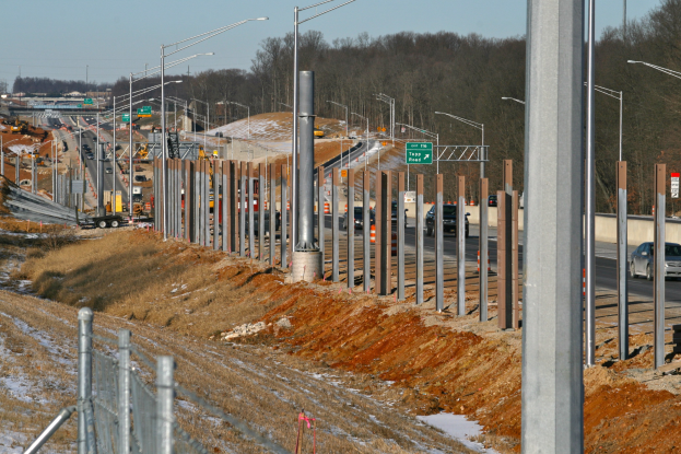 Baustelle mit Fahrzeugen, Pfählen, Laternen, Schildern, einem Zaun, Gras, Schnee, Bäumen und einem Himmel im Hintergrund.