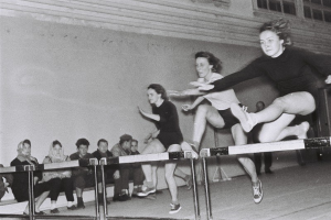 Zwei Frauen mitten im Sprung über ein Hindernis in einem Gymnastikraum, mit Zuschauern auf der linken Seite und einer fensterumgebenen Wand im Hintergrund.