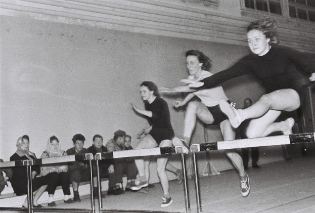 Zwei Frauen mitten im Sprung über ein Hindernis in einem Gymnastikraum, mit Zuschauern auf der linken Seite und einer fensterumgebenen Wand im Hintergrund.
