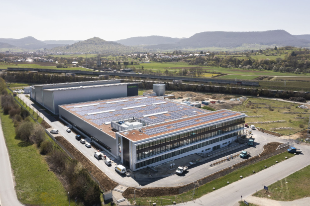 Luftaufnahme eines großen Gebäudes mit Solarmodulen auf dem Dach, umgeben von Fahrzeugen, Bäumen, Gras und Polen, mit Hügeln und einem klaren blauen Himmel im Hintergrund; das Gebäude ist Deutschlands erste solarbetriebene Fabrik.