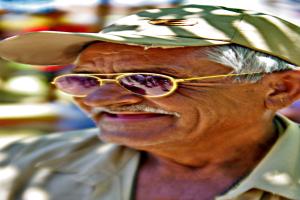 Ein alter Mann mit Hut und Brillengläsern, der fröhlich lächelt, mit einem unscharfen Hintergrund dahinter.