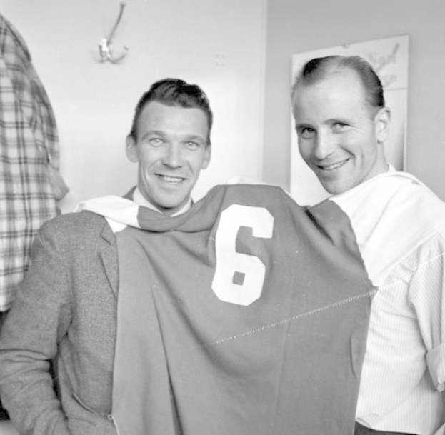 Zwei Männer in Schwarz-Weiß, die strahlend eine Fußballtrikot hochhalten, mit einer Tafel und einem Kleiderbügel an der Wand dahinter.