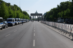 Eine Reihe von Polizeiwagen auf einer Stra√üe vor dem Brandenburger Tor in Berlin, Deutschland, mit Fahrradfahrern, F√ľ√üßg√§ngern, Barrieren, B√§umen und einem Bogen mit Statuen im Hintergrund.