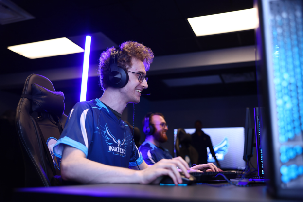 Ein Mann an einem Schreibtisch mit einem Computer, Headset und Brille, der ein Spiel in einem Raum mit einer anderen Person und Deckenbeleuchtung spielt.