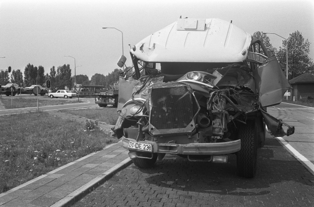 Ein Schwarz-Weiß-Foto eines schwer beschädigten Autos am Straßenrand mit eingedrückter Motorhaube und zersplitterten Fenstern, mit anderen Fahrzeugen, Laternenpfählen, Bäumen und einem Haus im Hintergrund.