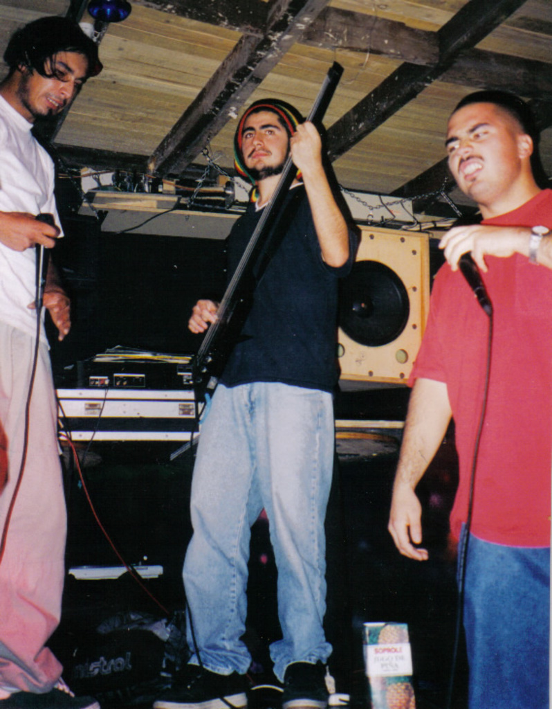 Ein Mann in schwarzem T-Shirt und blauen Jeans spielt Gitarre in der Mitte, flankiert von zwei Männern mit Mikrofonen, mit Lautsprechern und einem Verstärker auf einem Tisch dahinter.