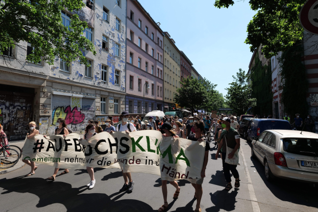 Eine Gruppe von Menschen marschiert auf einer Straße und hält ein Banner hoch, mit geparkten Autos auf der rechten Seite, einem Radfahrer auf der linken Seite und Gebäuden, Bäumen und einem Schild im Hintergrund.