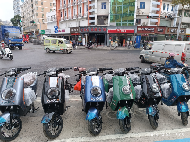 Eine Reihe von elektrischen Rollern, die an einer Straße geparkt sind, mit einer Person in einer Mücke davor, vor einem Hintergrund von Gebäuden, Verkehrsampeln, Bäumen und einem klaren Himmel.