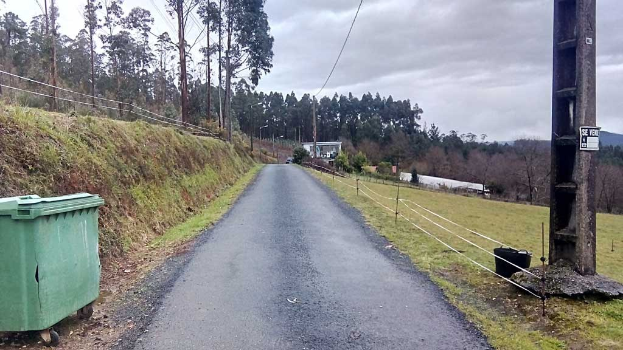 Grüner Müllcontainer auf der Seite einer Straße umgeben von Gras, Pflanzen, Bäumen, Polen, Dröhnen und einem Zaun unter einem bewölkten Himmel, mit einer Pfostenmontage auf der rechten Seite und verstreuten Gegenständen auf der linken Seite.