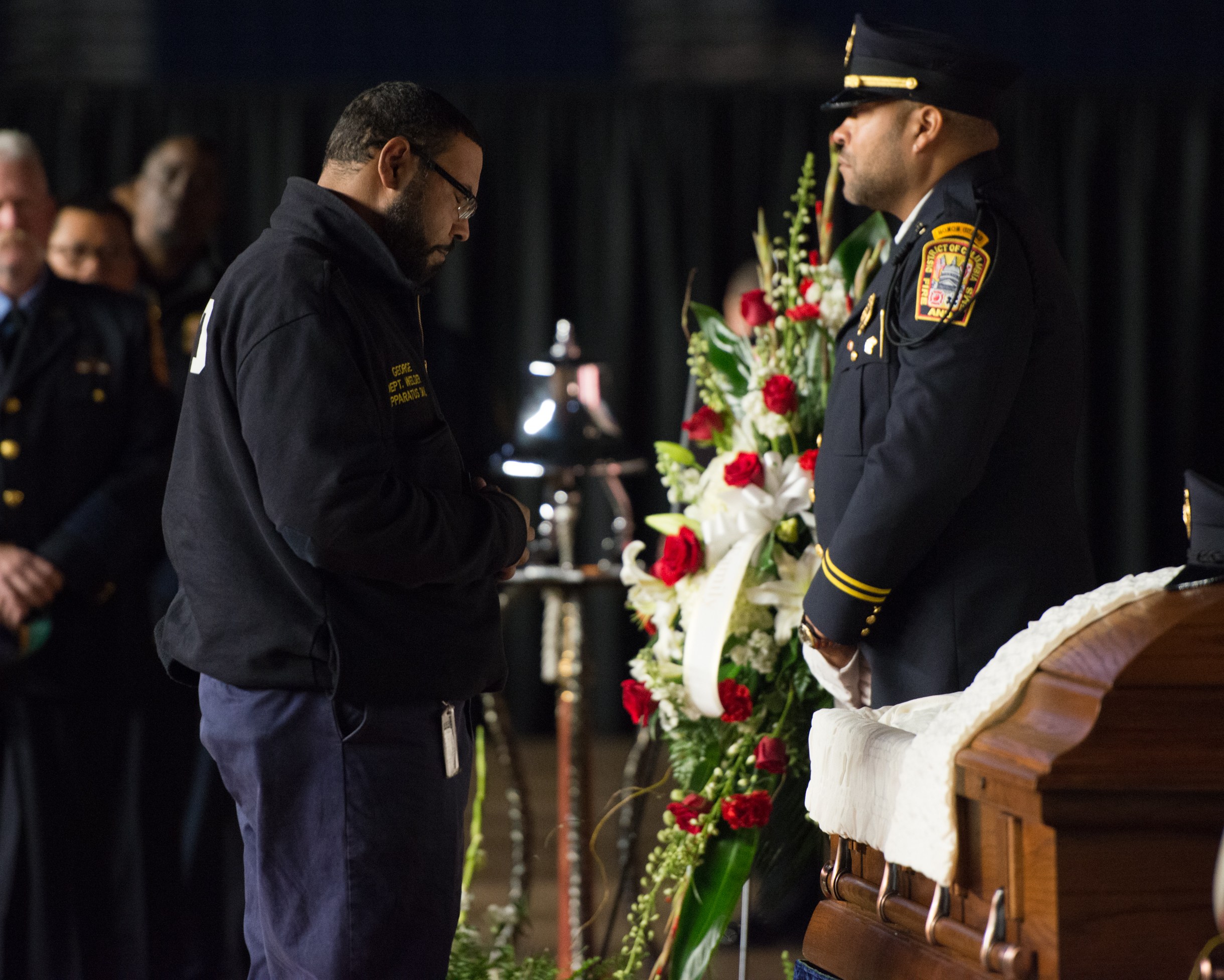 Zwei Polizeibeamte in Uniform stehen feierlich neben einem mit Blumen geschmückten Sarg, mit einigen Personen im Hintergrund.