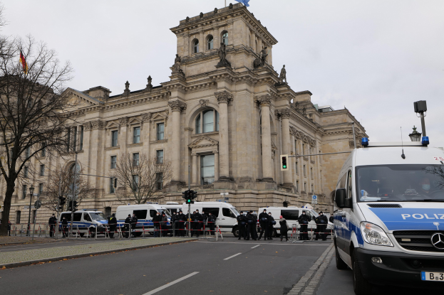 Eine Gruppe von Polizeibeamten vor dem Reichstaggebäude in Berlin, Deutschland, mit Fahrzeugen, einem Zaun, Verkehrszeichen, Laternen, Bäumen und Flaggen im Hintergrund.