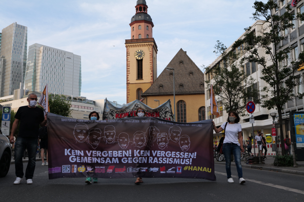 Eine Gruppe von Menschen in Masken, die eine Straße entlanggehen und ein Banner mit Text und Bildern halten, mit einem geparkten Auto links, Gebäuden, Bäumen, Schildern, Pfosten, einem Uhrenturm und einem klaren blauen Himmel im Hintergrund.