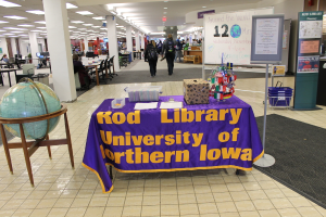 Eine Ausstellung mit einem Tisch, auf dem verschiedene Landesflaggen, ein Kasten, Papiere und ein Banner stehen, eine Kugel links daneben, ein Anschlagbrett rechts mit ein paar Leuten daneben und mehreren Tischen und Stühlen links.
