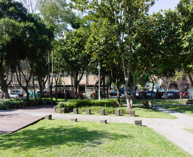 Ein Park mit grünem Gras, Bänken, Pflanzen, Bäumen, Laternenpfählen und Fahrzeugen auf einer Straße, mit Gebäuden und einem klaren blauen Himmel im Hintergrund.