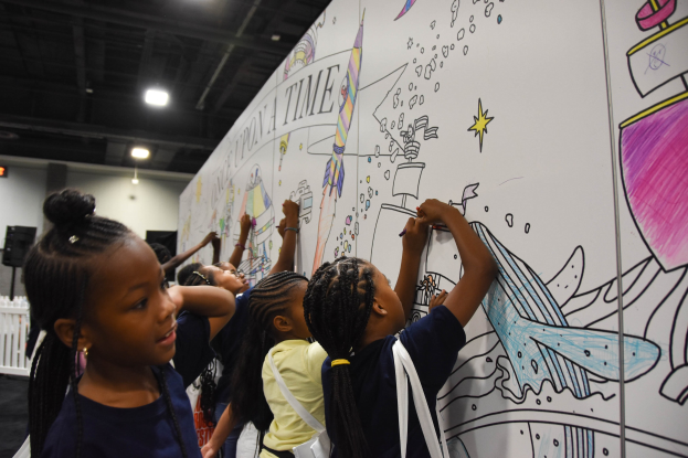Kinder malen auf einer großen Whiteboard an einer Indoor-Kunstausstellung, umgeben von Taschen, einem Zaun, Deckenlampen und Lautsprechern.