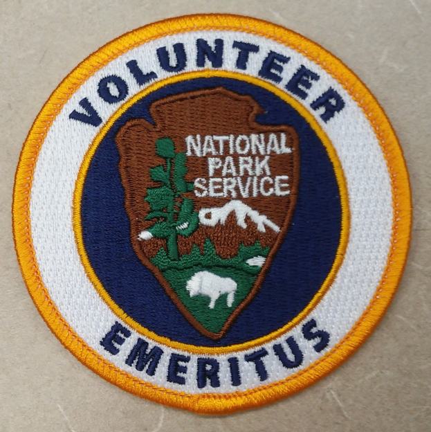 Runder Aufnäher mit blauem Rand und weißem Hintergrund, der schwarzen Text 'Volunteer National Park Service Emeritus' in einem gelben Kreis zeigt.