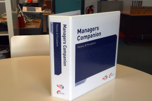 Ein Buch auf einem Tisch mit einem Stuhl dahinter, in der Nähe einer Tür, Feuerlöschern, Dateien und anderen Gegenständen.