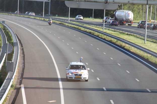 Ein Auto auf einer Autobahn mit einem stationären Polizeiwagen am Straßenrand, andere Fahrzeuge und Bäume, Geländer, Pfosten und Planken im Hintergrund.