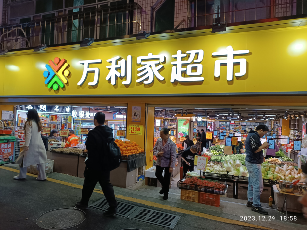 Eine Gruppe von Menschen geht an einem chinesischen Supermarkt in Hong Kong vorbei, dessen Schaufenster Gemüse, Obst und Schilder zeigt, sowie ein Mannloch auf der Straße.