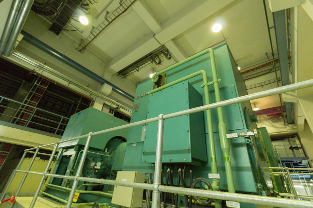 Großer industrieller Wärmetauscher in einem Gebäude, umgeben von Geländern, Rohren, Ventilen und Beleuchtungsgeräten.