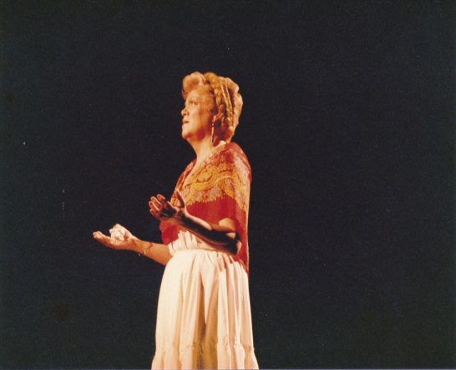 Eine Frau in einem weißen, hochgeschlossenen, langärmligen Kleid steht auf einer Bühne mit ausgebreiteten Armen, ihr Haar ist zu einem Dutt gebunden, vor einem dunklen Hintergrund.