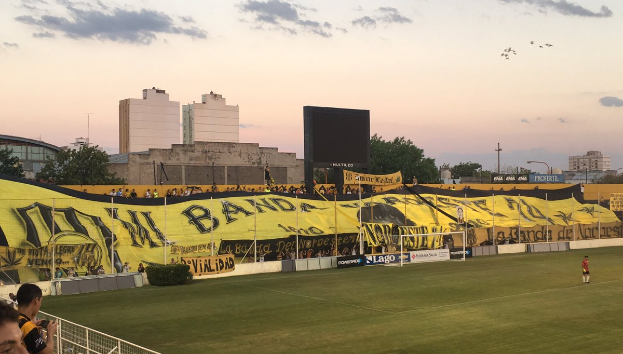 Ein Fußballfeld mit einer Gruppe von Menschen darauf, umgeben von einem Metallzaun, einem Torpfosten, einer Fahne mit der Aufschrift "Boca Juniors vs Real Sociedad", einigen Pflanzen, einem Anzeigebildschirm, Pfählen, Baumrinde, Bäumen, Gebäuden und einem bewölkten Himmel.