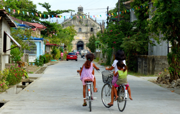 Drei Kinder fahren auf Fahrrädern eine von Bäumen gesäumte Straße entlang mit Gebäuden auf beiden Seiten, einem Auto und einem Gebäude im Hintergrund und Himmel darüber.