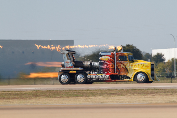 Ein Lkw mit Flammen, die aus der Hinterseite kommen, fahrt auf einer Stra├če, umgeben von Gras, einem Zaun, B├Ąumen, einem Geb├Ąude, einem Stra├čenpfahl, Dr├Ąhten und einem bew├Âlktem Himmel, mit einem sichtbaren Reifen und Rad.