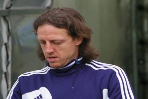 Ein Mann mit langen Haaren in einem blauen T-Shirt steht ernst vor einer grünen Wand und sieht aus wie ein Trainer.