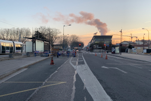 Eine Straßenszene in einer Stadt mit Verkehrskegeln, einem Bus, Laternen, Bäumen, Gebäuden, Kränen und Rauch, der aus einem entfernten Gebäude aufklarendem Himmel aufsteigt.