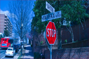 Eine Straßenszene mit mehreren Gebäuden, Bäumen, geparkten Autos, einem Bus, Schildern an Masten und einem blauen bewölktem Himmel, mit einer Mastlaterne.
