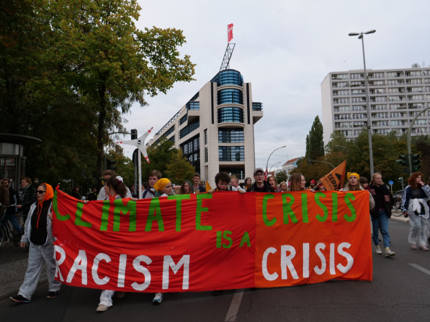 Eine Gruppe von Menschen geht eine Straße entlang und hält ein Banner mit der Aufschrift "Klima-Krise ist eine Krise", mit Bäumen, Laternenmasten und Gebäuden am Straßenrand und dem Himmel im Hintergrund.