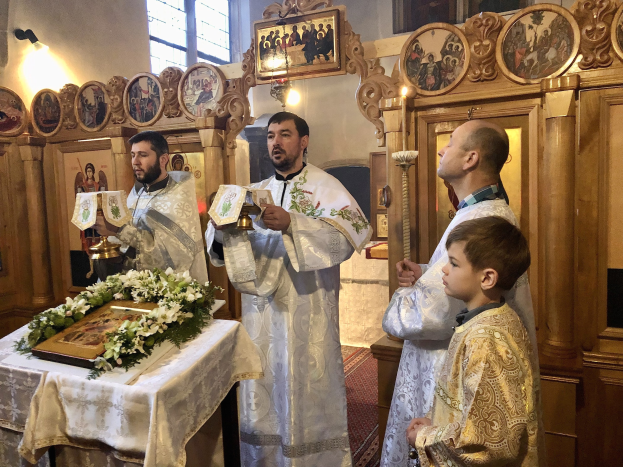 Eine Gruppe von Menschen um einen Tisch in einer Kirche herumstehend, mit Büchern haltend, mit einem Tuch und einem Blumenstraß auf dem Tisch, und Foto-Rahmen, Lichtern, einem Fenster und einem hölzernen Gegenstand im Hintergrund.