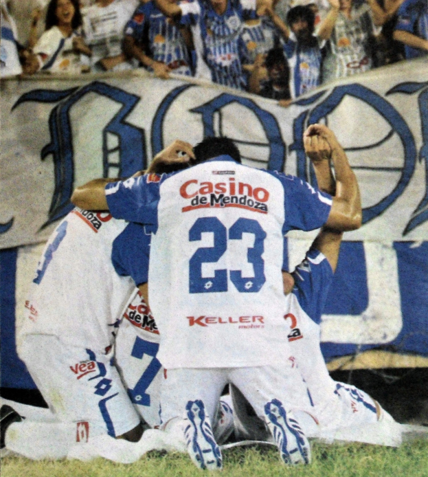 Eine Gruppe von Fußballspielern feiert ein Tor auf dem Feld mit einer jubelnden Menge im Hintergrund und einem Banner mit Text dahinter.